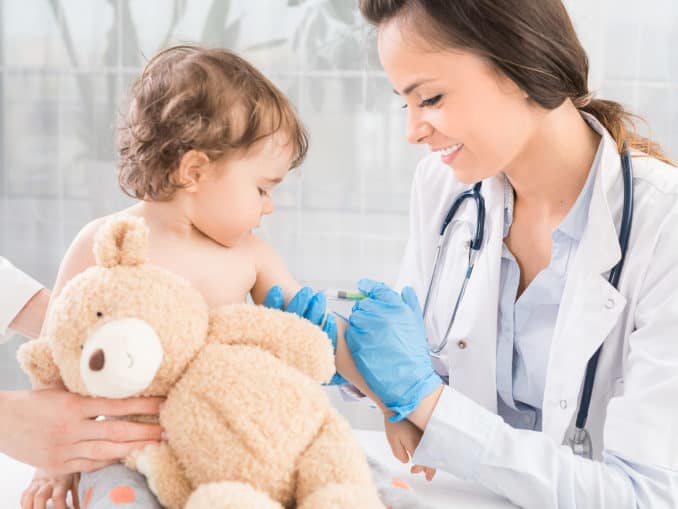 Freiwillige gesetzliche Versicherung für Kinder: Eine gute Lösung? 1 Junge Frau Kinderarztin führt eine Impfung eines kleinen Mädchens durch. Das Mädchen hält ein Maskottchen.